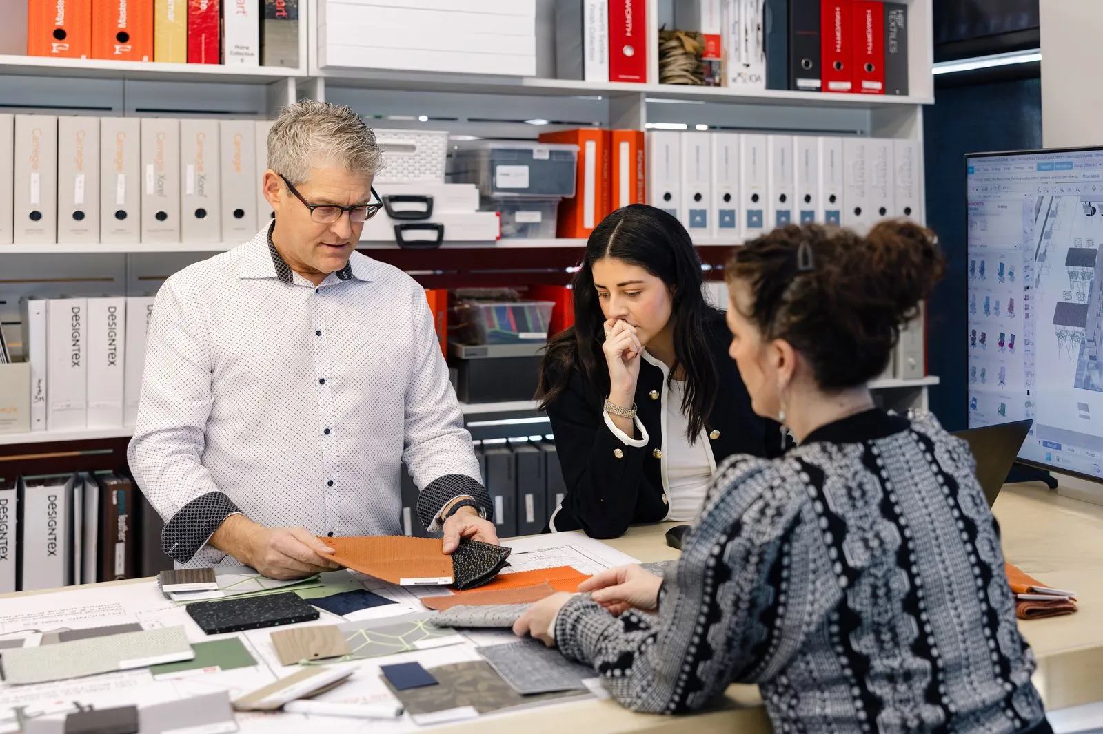 Three people reviewing finish samples and floor plans at a table in front of shelves of manufacturer binders